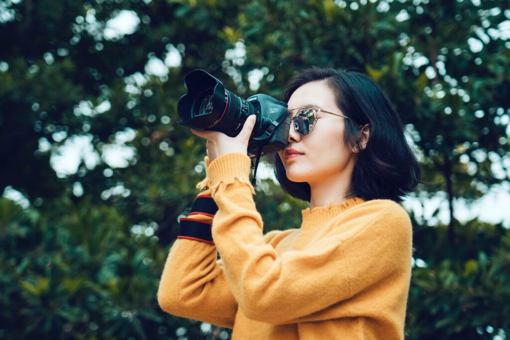 This is an image of an Asian woman with short black hair wearing a yellow sweater holding a Canon camera to her eye