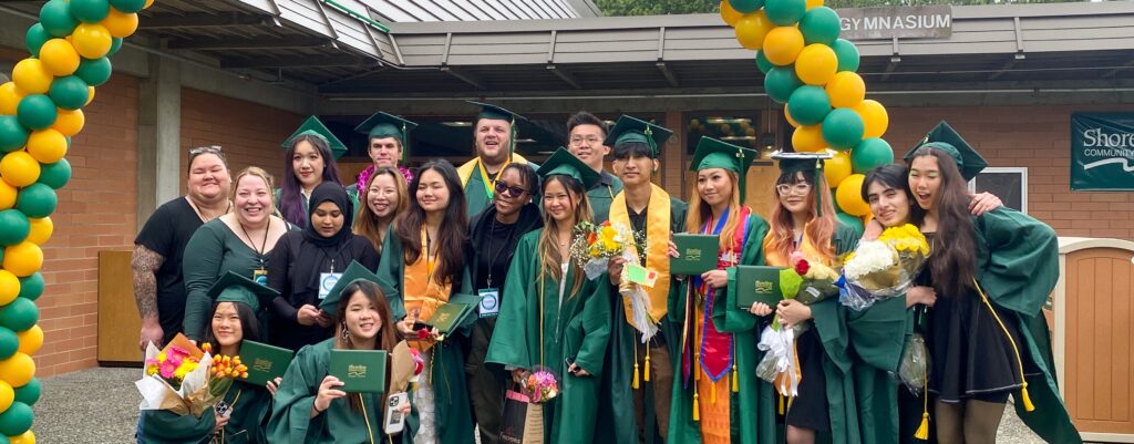 Group photo of smiling graduates standing beneath an arch of green and yellow balloons. 
