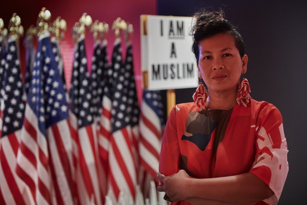 This is an image of the speaker in front of a row of flags with a sign that says "I am a muslim" in the background.