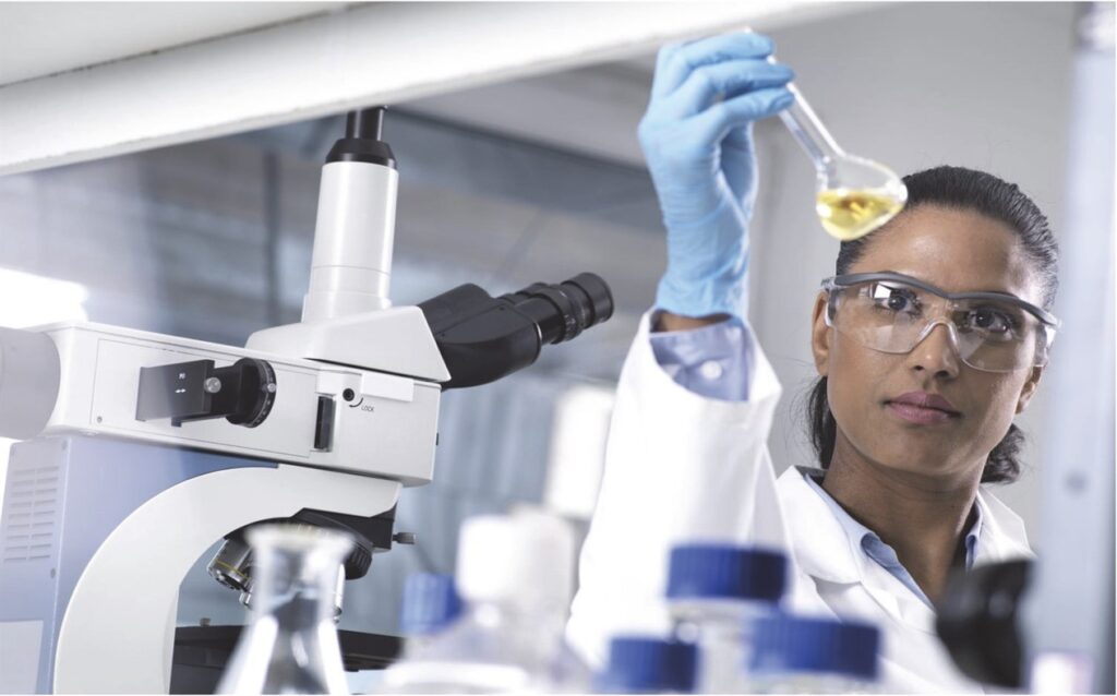 This is a woman dressed in a lab coat in front of a microscope holding a beaker with yellow liquid.