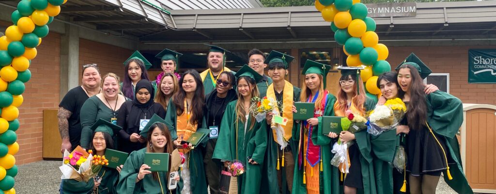 A group of smiling graduates gathered under an archway of green and yellow balloons.