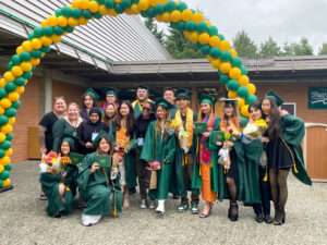 Group of smiling graduates under an arch of green and yellow balloons.