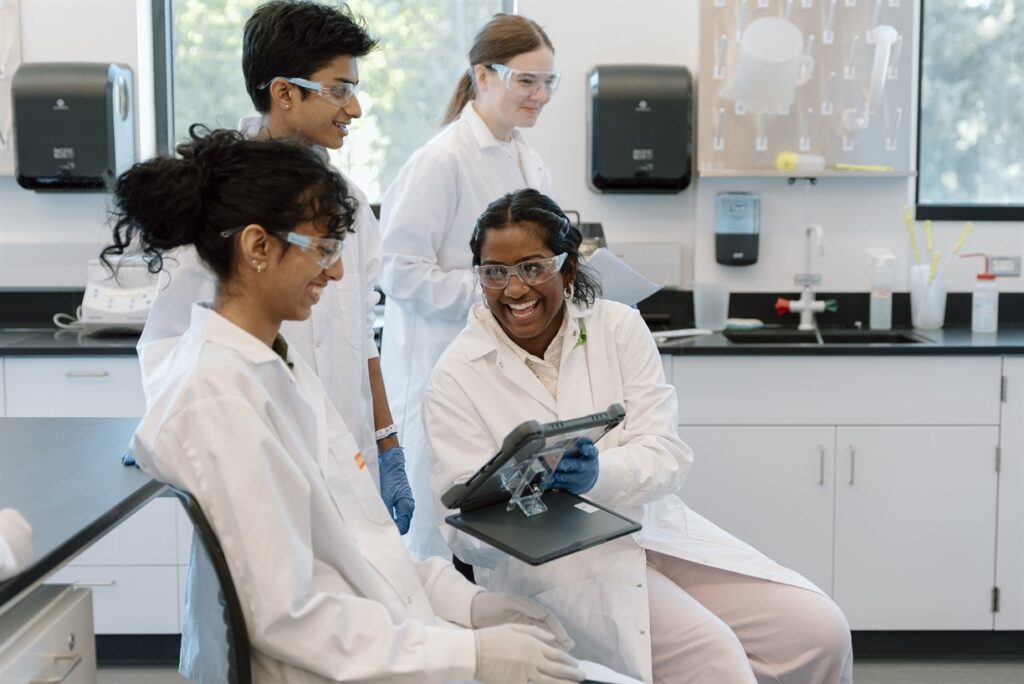 Image of smiling students in a science lab wearing lab coats. 